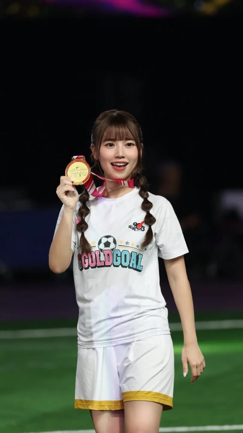 《足球女將》成員「表妹」吳梓清(Mona)身穿 GOLDGOAL 白色球衣,在球場上微笑拿著金牌展示。