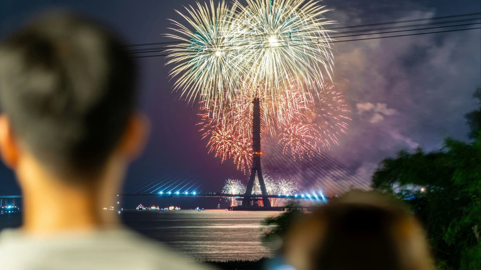 夜晚海邊看煙火的視角,前景是兩位觀賞者的背影模糊輪廓,中景為一座大型斜張橋,背景夜空中綻放著金色與紅色的絢麗煙火,水面上映照著煙火倒影。