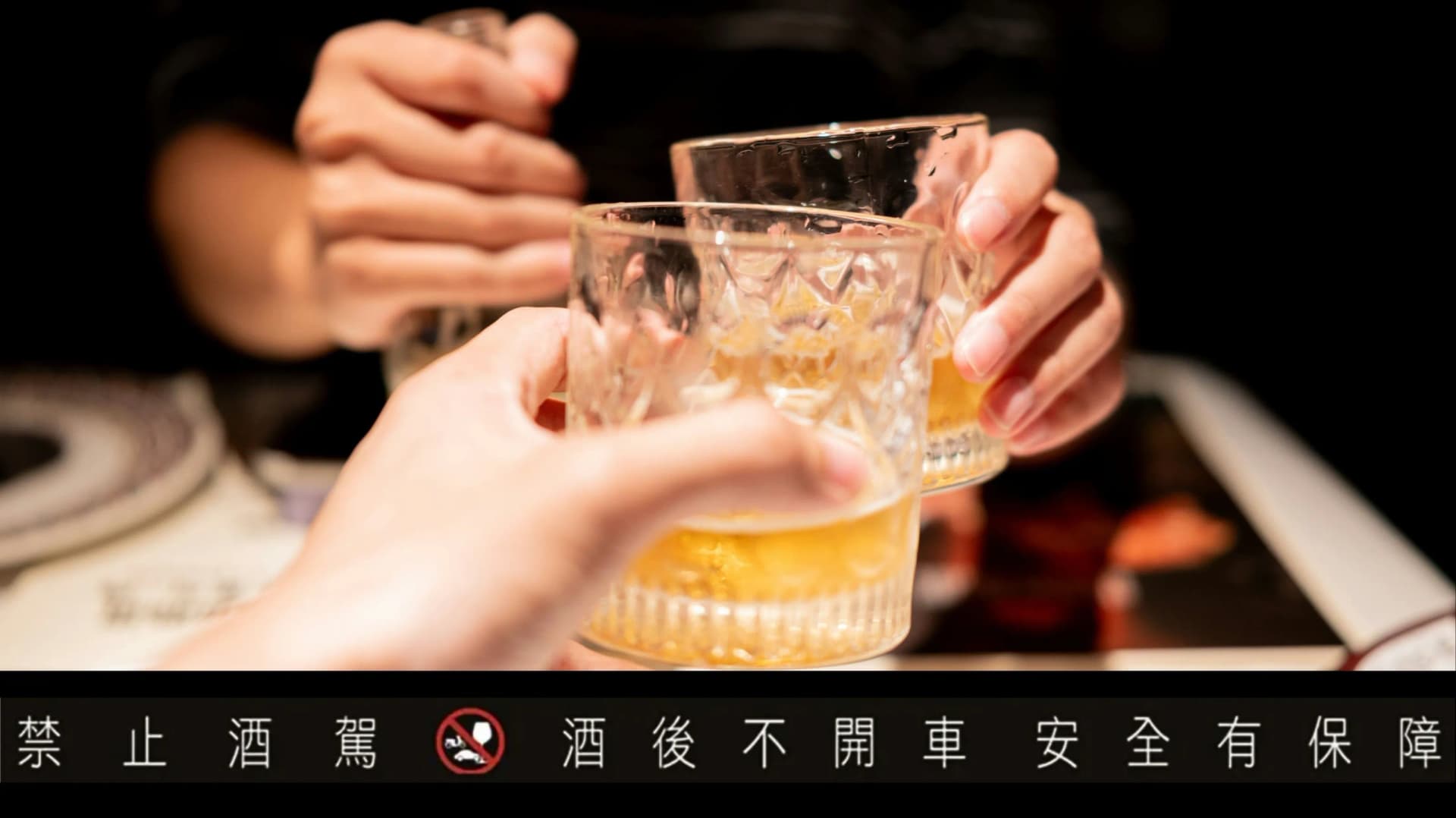 在暗色調的餐廳或酒吧中,兩手拿著裝有金黃色酒液的玻璃杯碰撞乾杯的特寫鏡頭。圖片下方標有黑底白字的警語「禁止酒駕 酒後不開車 安全有保障」,中央附有禁止酒駕的圖示。
