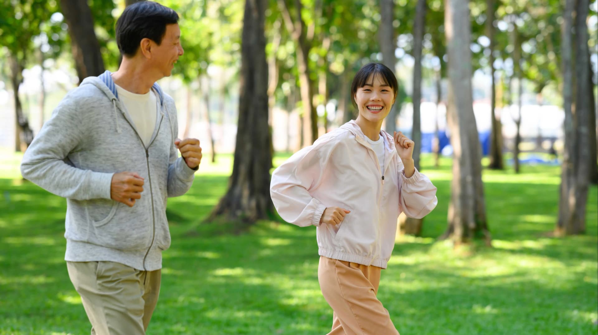 一對面帶著微笑的男女在綠樹成蔭的公園慢跑,陽光灑落,女子身穿淺粉色外套和蜜桃色長褲,燦爛地笑著。男子則穿著灰色連帽衫和卡其色長褲。