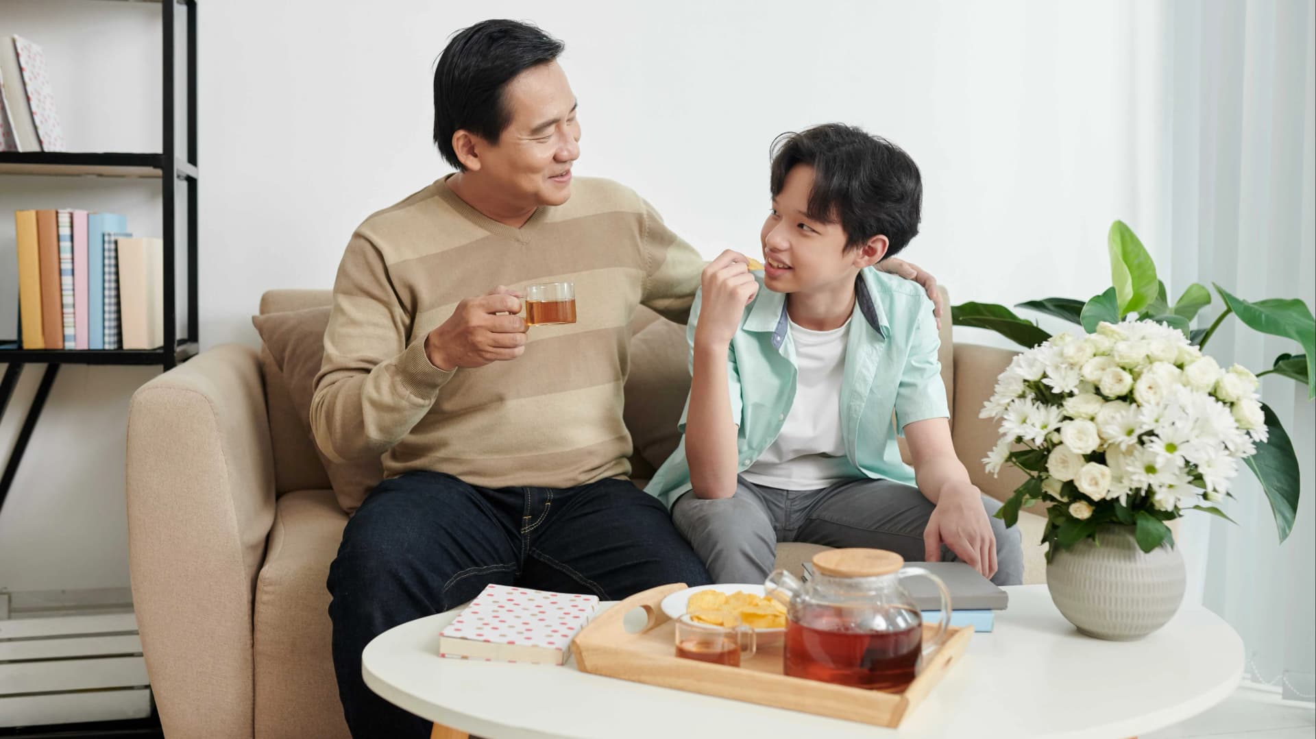 一位成年人和一位青少年坐在沙發上,成年人一隻手臂搭在青少年的肩上,兩人前方的桌子上擺放著一些飲料、小吃和一瓶白色鮮花,後方有書架與書本。