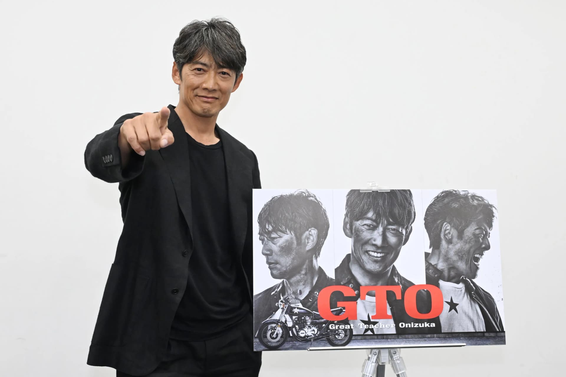 日籍男神反町隆史宣傳經典特別篇戲劇作品現場景致。畫面呈現反町隆史身穿黑色西裝手指向前方,身旁放置三款經典造型宣傳海報,展現型男魅力風采景致。