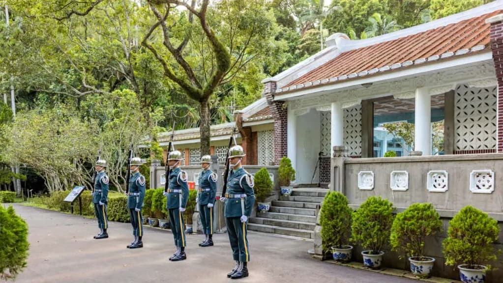 桃園景點慈湖陵寢儀隊交接現場景致。畫面呈現三軍儀隊士兵身穿制服於閩南式黑瓦白牆建築前站崗,周圍環繞綠意盎然樹木,展現莊嚴歷史氛圍與景觀風貌景致。