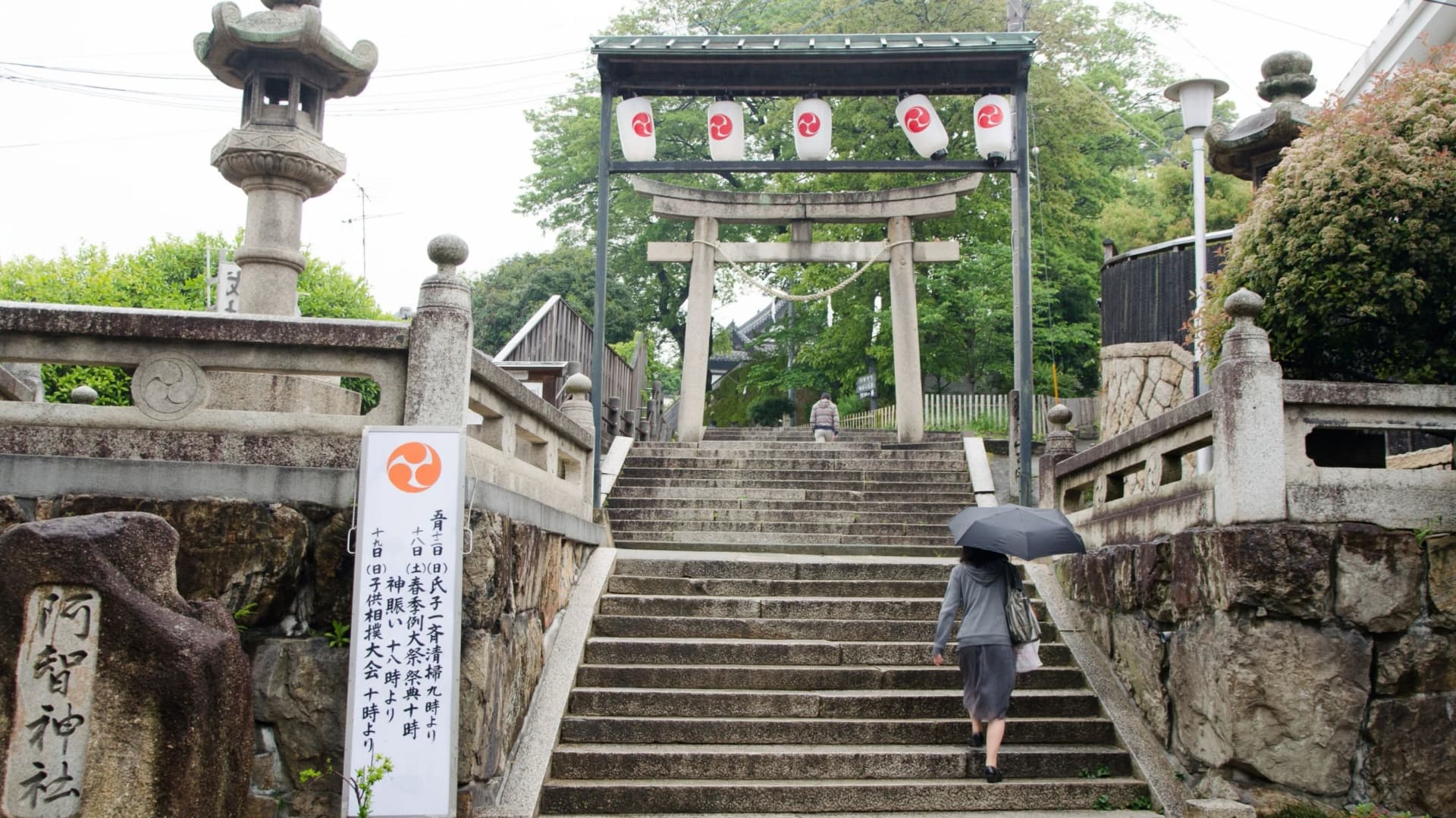 岡山倉敷阿智神社入口處,參拜者撐傘走上高聳的石階梯,前方立有鳥居與刻有「阿智神社」的石碑。